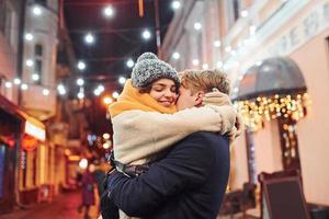 Pareja joven positiva en ropa de abrigo abrazándose unos a otros en la calle decorada de Navidad foto