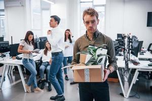 hombre elegante con gafas sosteniendo una caja con una planta frente a sus colegas en la oficina foto