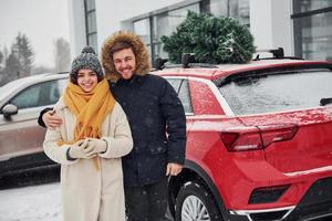 Young couple standing near car with tree on the top. Together outdoors at winter time photo