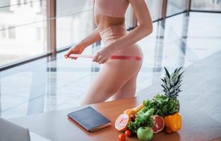 Holding measuring tape. Woman with slim body type and clean skin is indoors in sportive wear photo