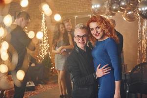 Young couple standing together against their friends in christmas decorated room and celebrating New Year photo