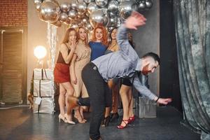 Young women in festive clothes standing indoors of christmas decorated room together and looks at man that have fun doing tricks photo