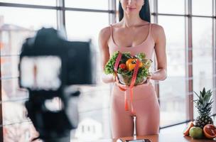 joven vlogger de fitness haciendo video en interiores parándose cerca de la mesa con comida saludable foto