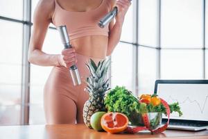 Female athlete in sportive wear holds dumbbells and standing near table with laptop and healthy food photo