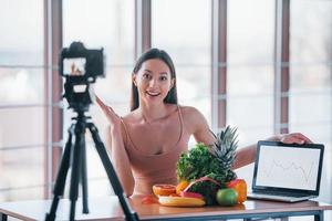 joven vlogger de fitness haciendo video en interiores sentado cerca de la mesa con comida saludable foto