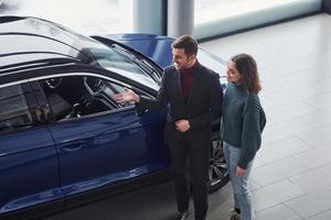 Professional salesman assisting young girl by choosing new modern automobile indoors photo