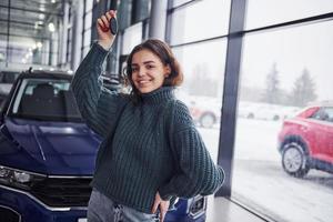 una joven alegre sostiene las llaves y se para frente a un auto nuevo y moderno en el interior foto