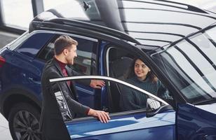 Professional salesman assisting young girl by choosing new modern automobile indoors photo