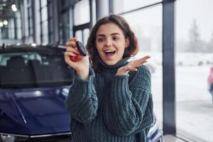 Cheerful young woman holds keys and standing in front of modern new car indoors photo