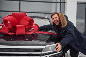 Happy owner of new modern car standing outdoors and smiling. Red gift length on the automobile photo