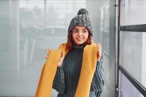 hermosa chica alegre con bufanda amarilla y ropa abrigada de pie en el interior contra el fondo con reflejos foto