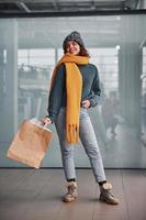 Beautiful cheerful girl in yellow scarf and in warm clothes standing indoors with shopping bags in hands photo