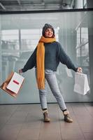 hermosa chica alegre con bufanda amarilla y ropa abrigada de pie en el interior con bolsas de compras en las manos foto