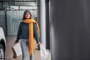 hermosa chica alegre con bufanda amarilla y ropa abrigada de pie en el interior con bolsas de compras en las manos cerca del auto foto