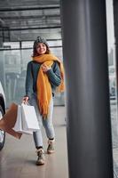 hermosa chica alegre con bufanda amarilla y ropa abrigada de pie en el interior con bolsas de compras en las manos foto