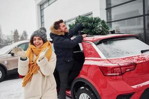 Young couple standing near car with tree on the top. Together outdoors at winter time photo