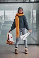 Beautiful cheerful girl in yellow scarf and in warm clothes standing indoors with shopping bags in hands photo