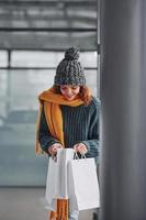 Beautiful cheerful girl in yellow scarf and in warm clothes standing indoors with shopping bags in hands photo