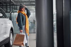 hermosa chica alegre con bufanda amarilla y ropa abrigada de pie en el interior con bolsas de compras en las manos cerca del auto foto