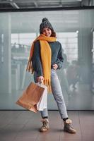 Beautiful cheerful girl in yellow scarf and in warm clothes standing indoors with shopping bags in hands photo