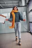 Beautiful cheerful girl in yellow scarf and in warm clothes standing indoors with shopping bags in hands photo