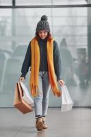 Beautiful cheerful girl in yellow scarf and in warm clothes standing indoors with shopping bags in hands photo