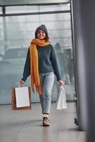 Beautiful cheerful girl in yellow scarf and in warm clothes standing indoors with shopping bags in hands photo