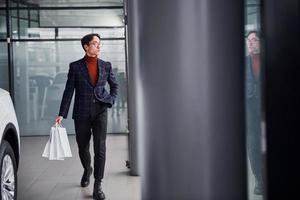 Holding shopping bags. Young business man in luxury suit and formal clothes is indoors near the car photo