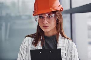 retrato de una joven trabajadora con casco protector, anteojos y bloc de notas. de pie en la oficina foto