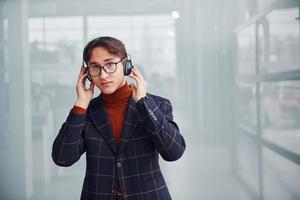 Listening to the music in headphones. Young business man in luxury suit and formal clothes is indoors in the office photo