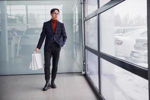 With shopping bags. Young business man in luxury suit and formal clothes is indoors in the office photo