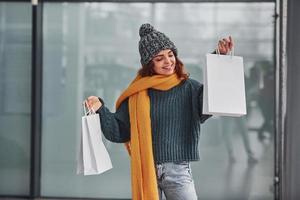 Beautiful cheerful girl in yellow scarf and in warm clothes standing indoors with shopping bags in hands photo