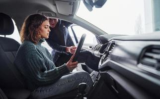 Professional salesman assisting young girl by choosing new modern automobile indoors. Woman sitting inside photo