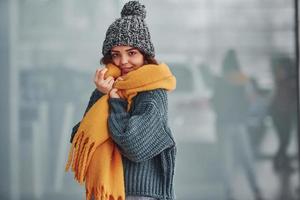 Beautiful cheerful girl in yellow scarf and in warm clothes standing indoors against background with reflections photo