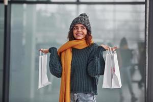 Smiling and feeling happy. Beautiful cheerful girl in yellow scarf and in warm clothes standing indoors with shopping bags in hands photo