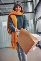 Beautiful cheerful girl in yellow scarf and in warm clothes standing indoors with shopping bags in hands photo