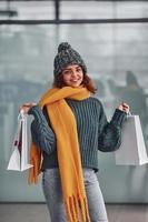 Smiling and feeling happy. Beautiful cheerful girl in yellow scarf and in warm clothes standing indoors with shopping bags in hands photo