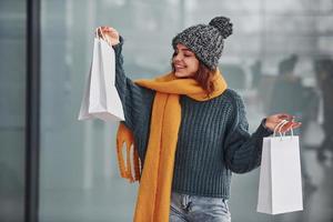 Smiling and feeling happy. Beautiful cheerful girl in yellow scarf and in warm clothes standing indoors with shopping bags in hands photo