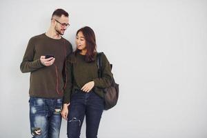 Cheerful multi ethnic couple with backpack and phone standing together indoors in the studio against white background photo
