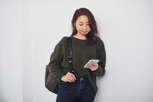 Portrait of happy asian young girl with backpack and phone that standing indoors in the studio against white background photo