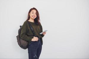 Portrait of happy asian young girl with backpack and phone that standing indoors in the studio against white background photo