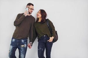 Cheerful multi ethnic couple with backpack and phone standing together indoors in the studio against white background photo