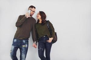 Cheerful multi ethnic couple with backpack and phone standing together indoors in the studio against white background photo