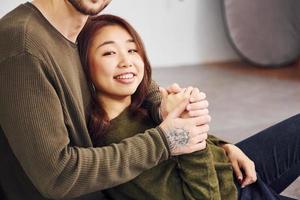 Happy multi ethnic couple in casual clothes sitting together indoors in the studio. Caucasian guy with asian girlfriend photo