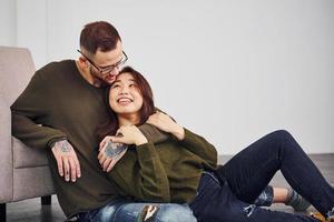 Happy multi ethnic couple in casual clothes sitting together indoors in the studio. Caucasian guy with asian girlfriend photo