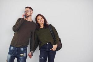 Cheerful multi ethnic couple with backpack and phone standing together indoors in the studio against white background photo