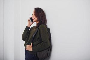 Portrait of happy asian young girl with backpack and phone that standing indoors in the studio against white background photo