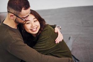 Happy multi ethnic couple in casual clothes sitting together indoors in the studio. Caucasian guy with asian girlfriend photo