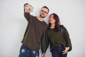 alegre pareja multiétnica con ropa informal haciendo selfie en el interior del estudio con fondo blanco foto