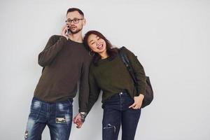 alegre pareja multiétnica con mochila y teléfono de pie juntos en el interior del estudio con fondo blanco foto